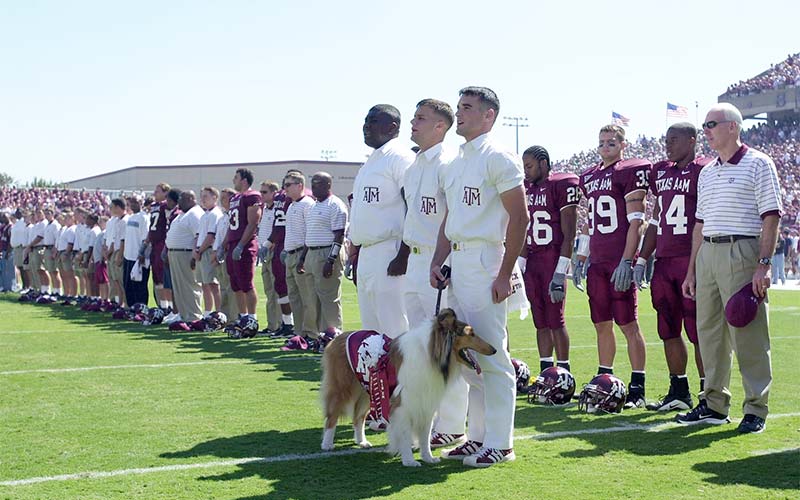 Coaches, players and students on the football field with a dog in a Aggies covering